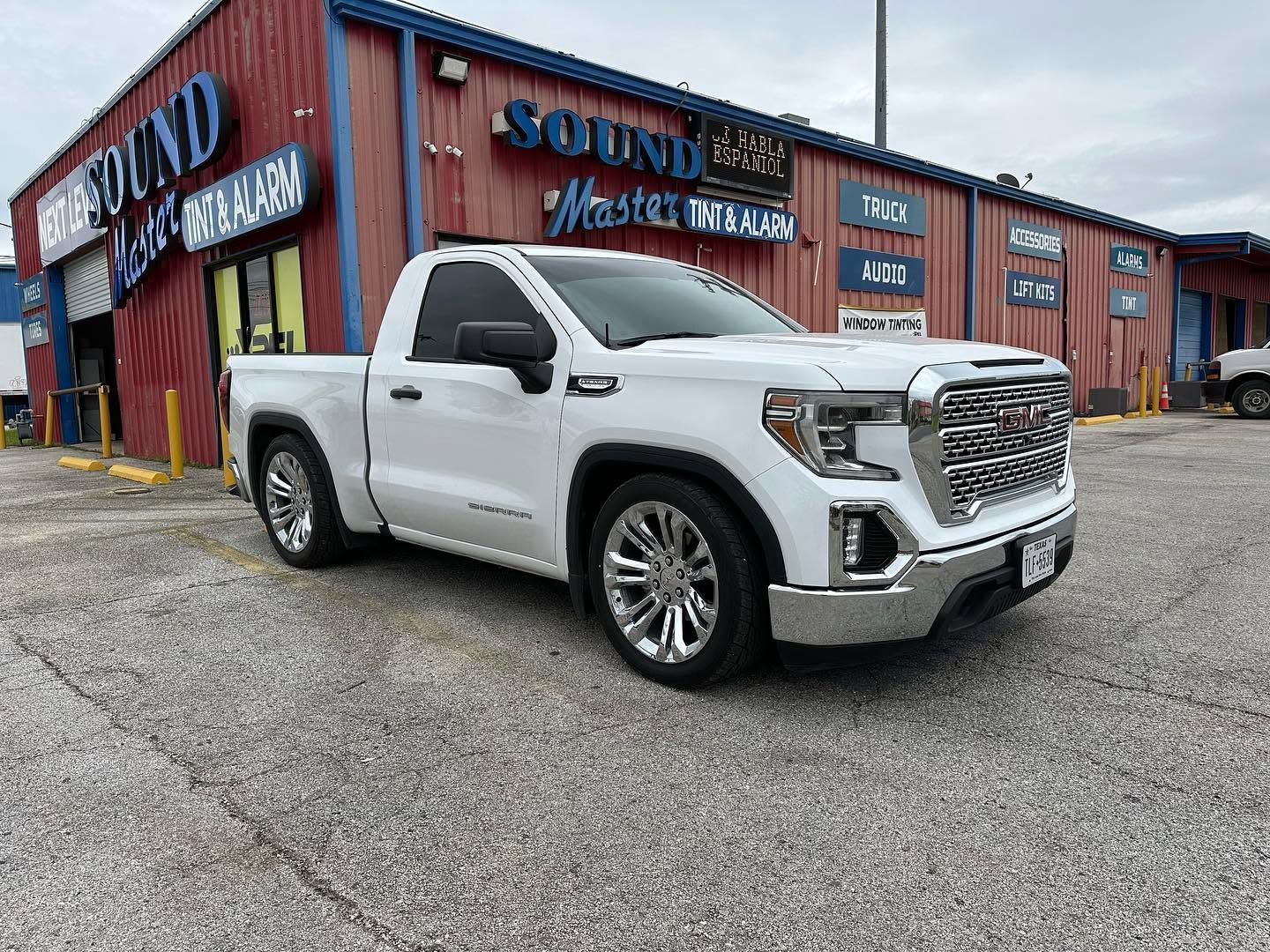 White GMC pickup truck parked outside a red brick auto dealership with a blue sign.