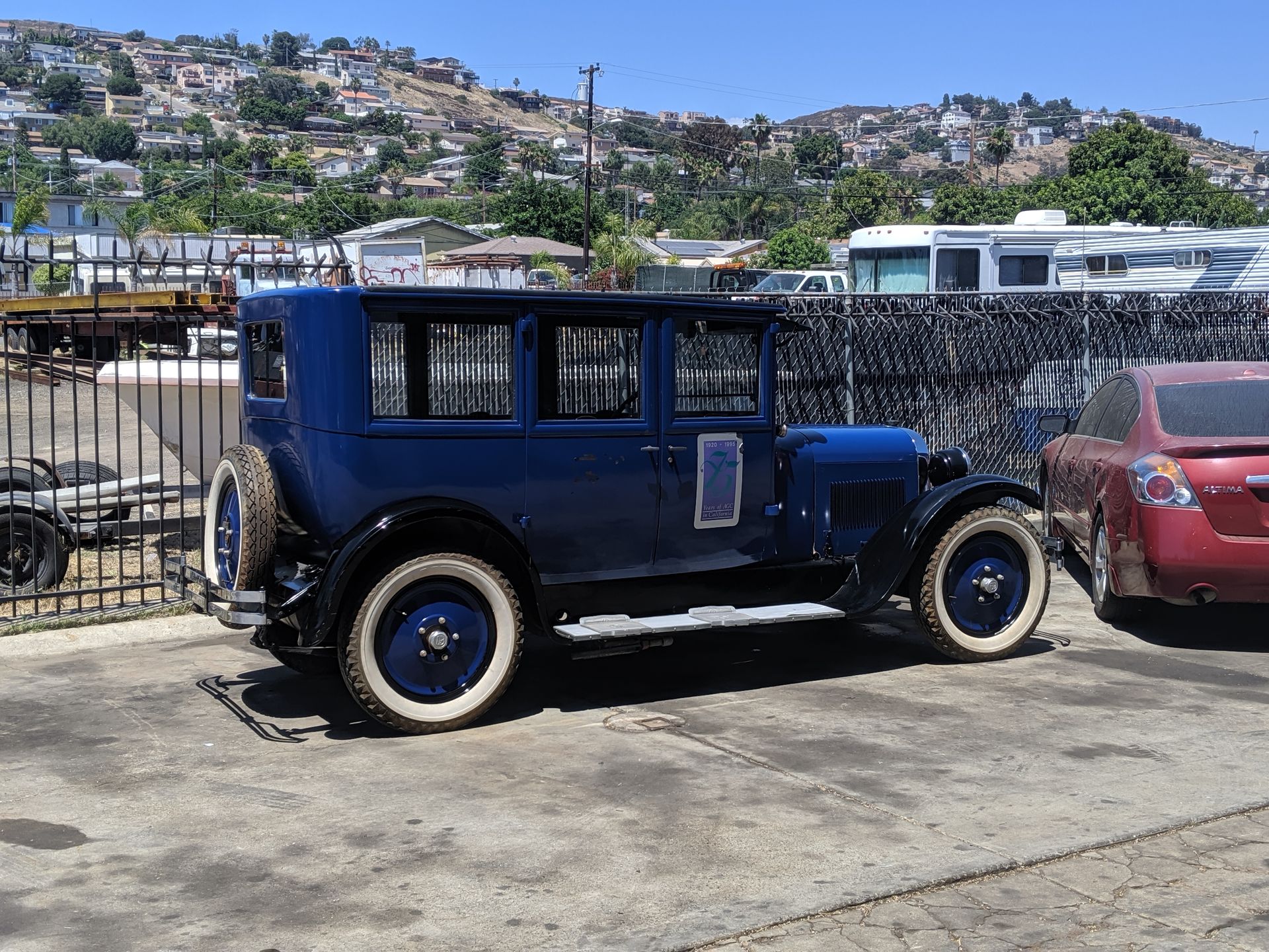 Blue vintage car parked beside a red car on a sunny lot with hillside houses in the background