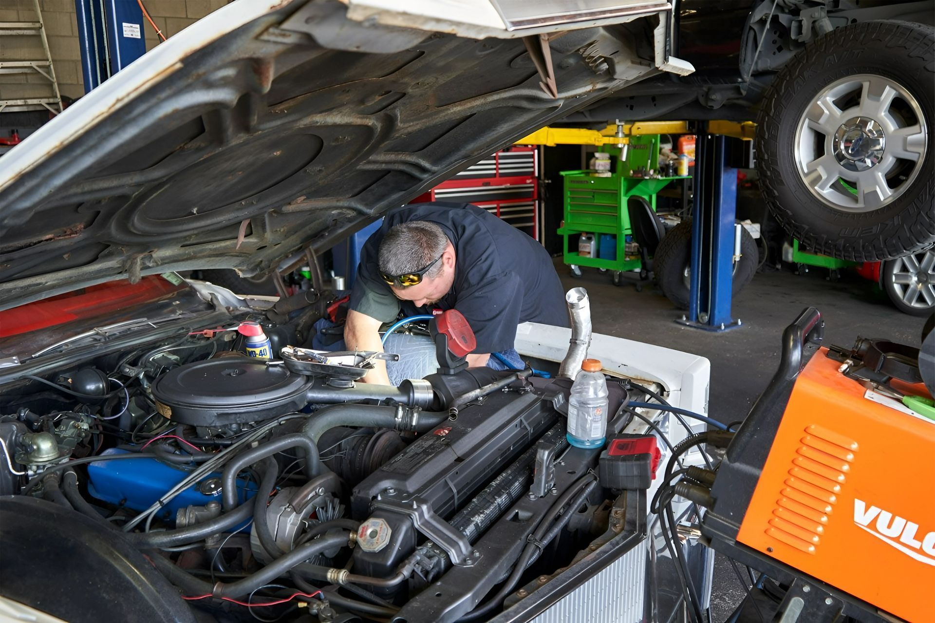 Mechanic working under a raised car hood in an auto repair shop with tools and equipment nearby