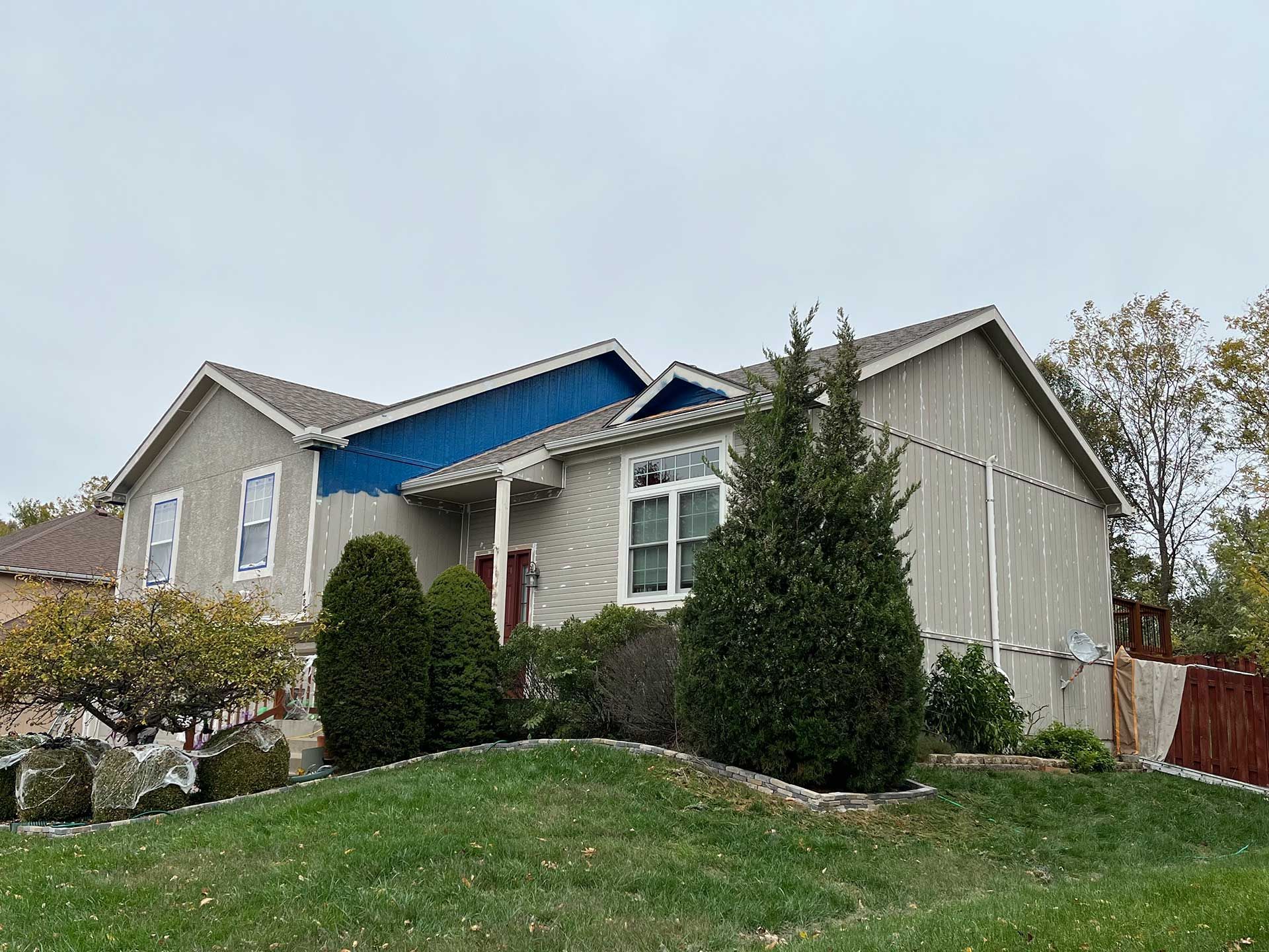 a house being painted blue, green lawn, bushes, and overcast sky