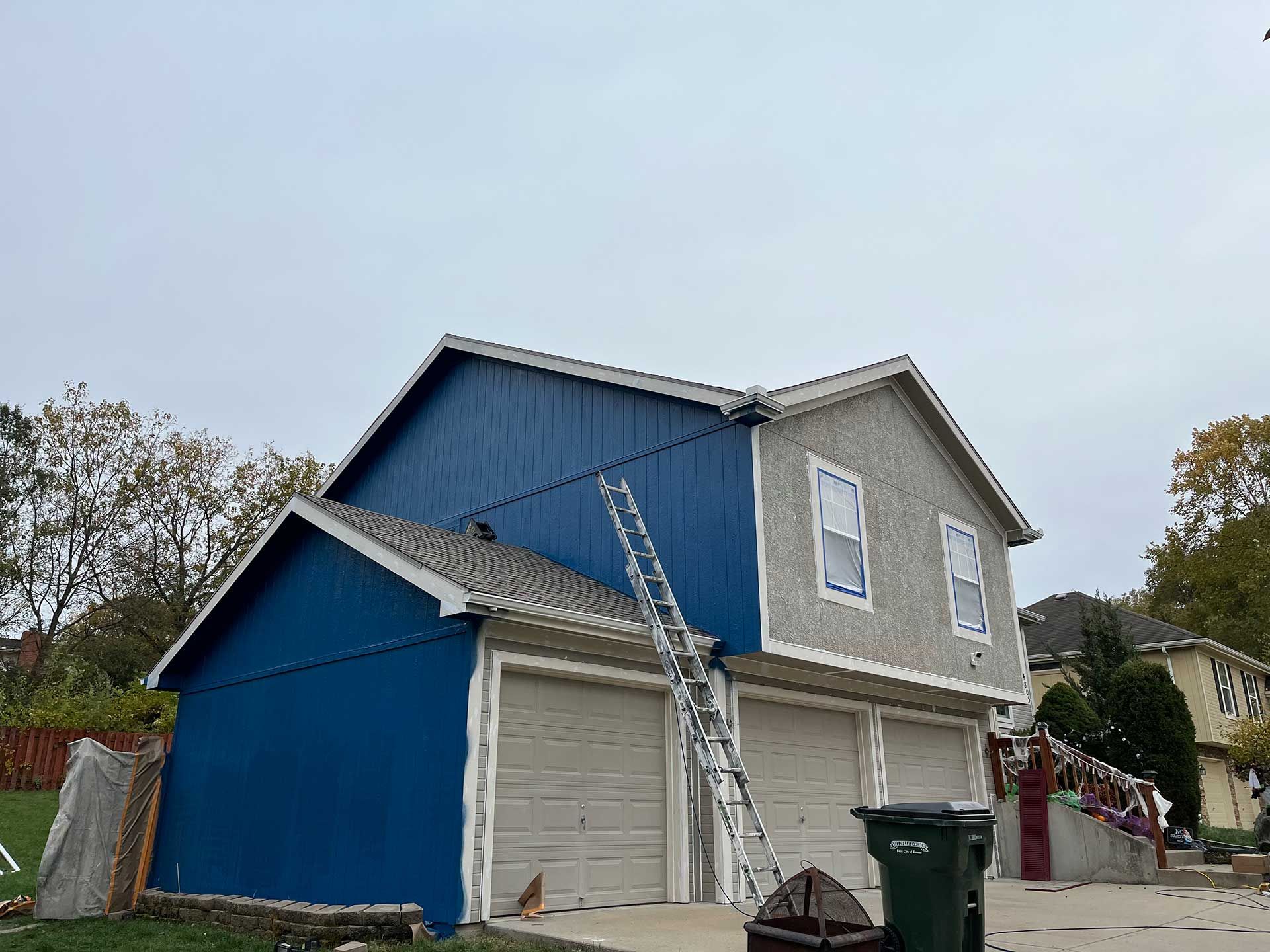 two-story house with blue and gray siding; ladder leans against the building near garage doors