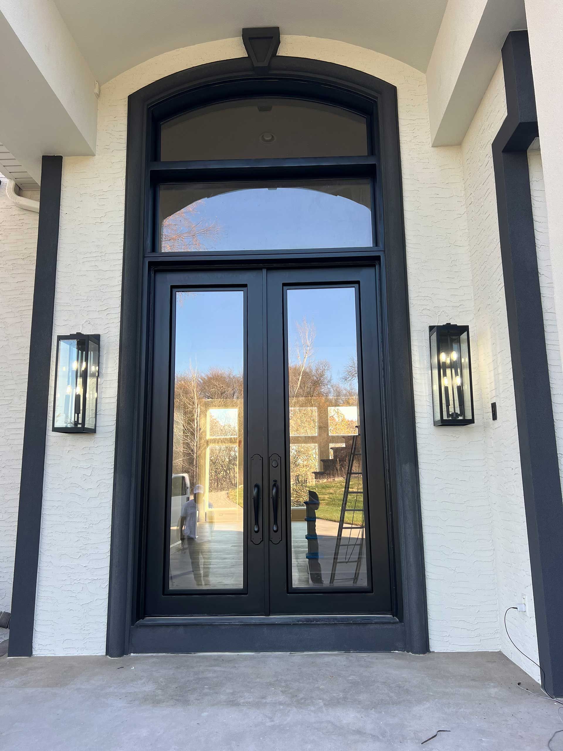 black double doors with arched transom window, flanked by sconces on white stucco facade