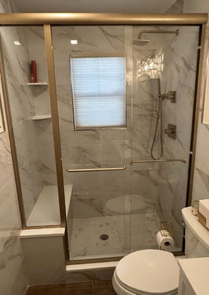 Modern bathroom with marble tile, glass shower door, and gold trim. Includes a toilet, window, and built-in bench.