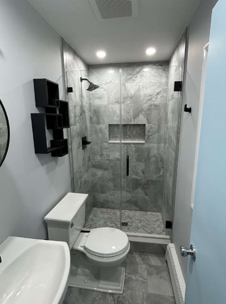 Modern bathroom with gray tiled shower, toilet, floating shelves, and sink.