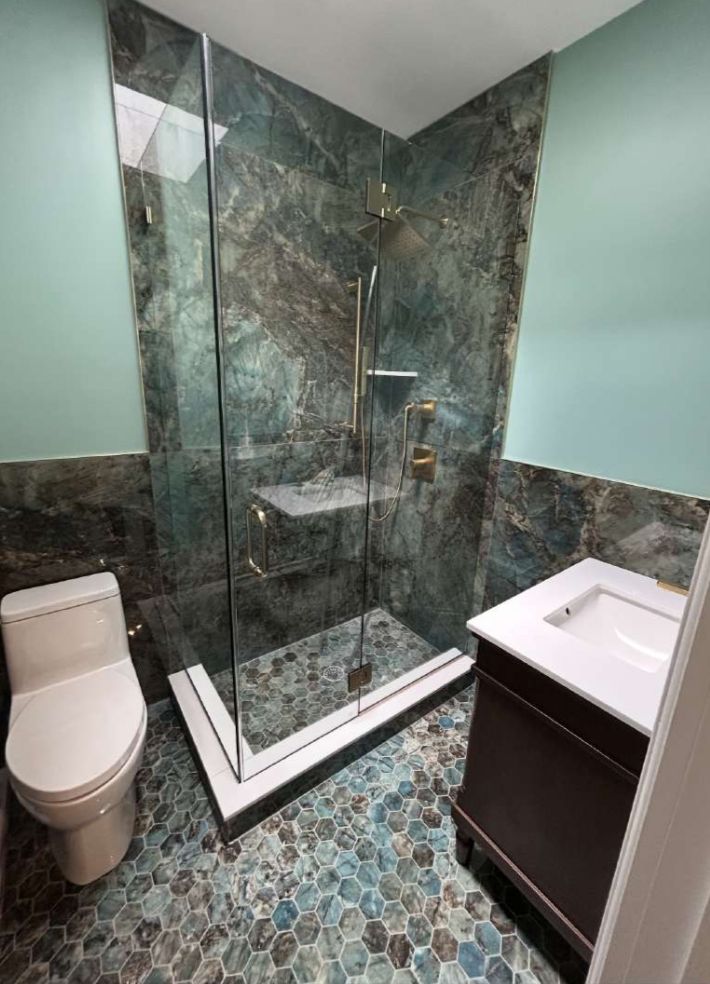 Modern bathroom with teal stone tile shower, toilet, and dark vanity. Blue and teal pebble floor.