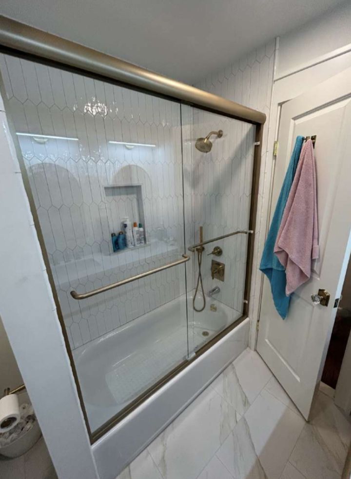 Shower with glass doors, gold frame, built-in shelf, white tile walls, and a partially open white door with towels.