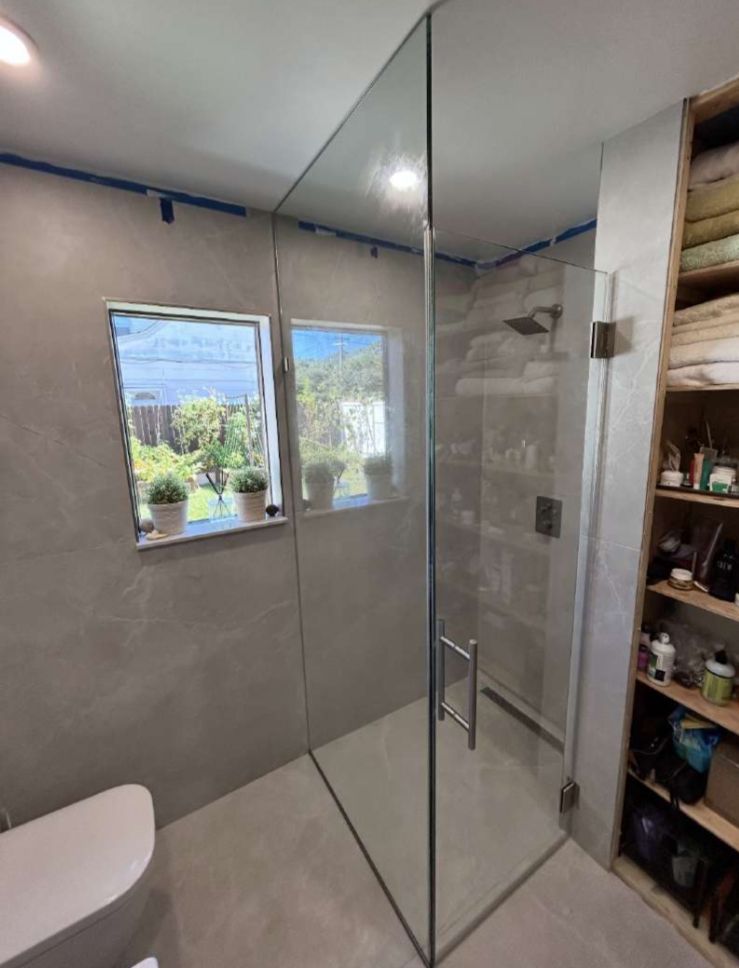 Modern bathroom with glass shower enclosure, two windows, and open shelving. Gray walls and flooring.