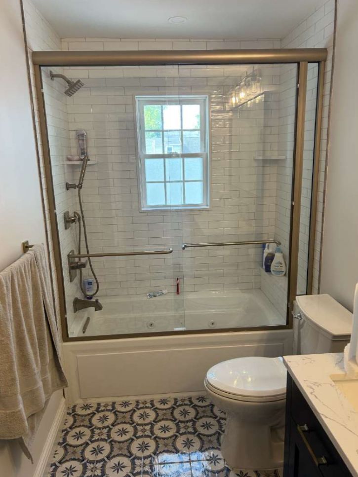 Bathroom with white subway tile, gold-framed glass shower, patterned tile floor, toilet, and window.