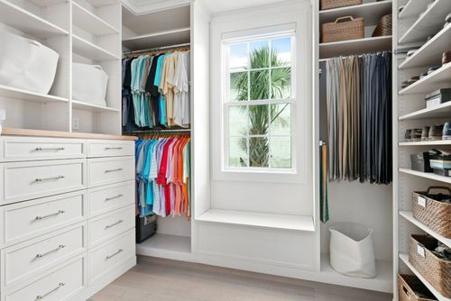 Bright walk-in closet with white shelves, drawers, hanging clothes, and a window seat by a central window