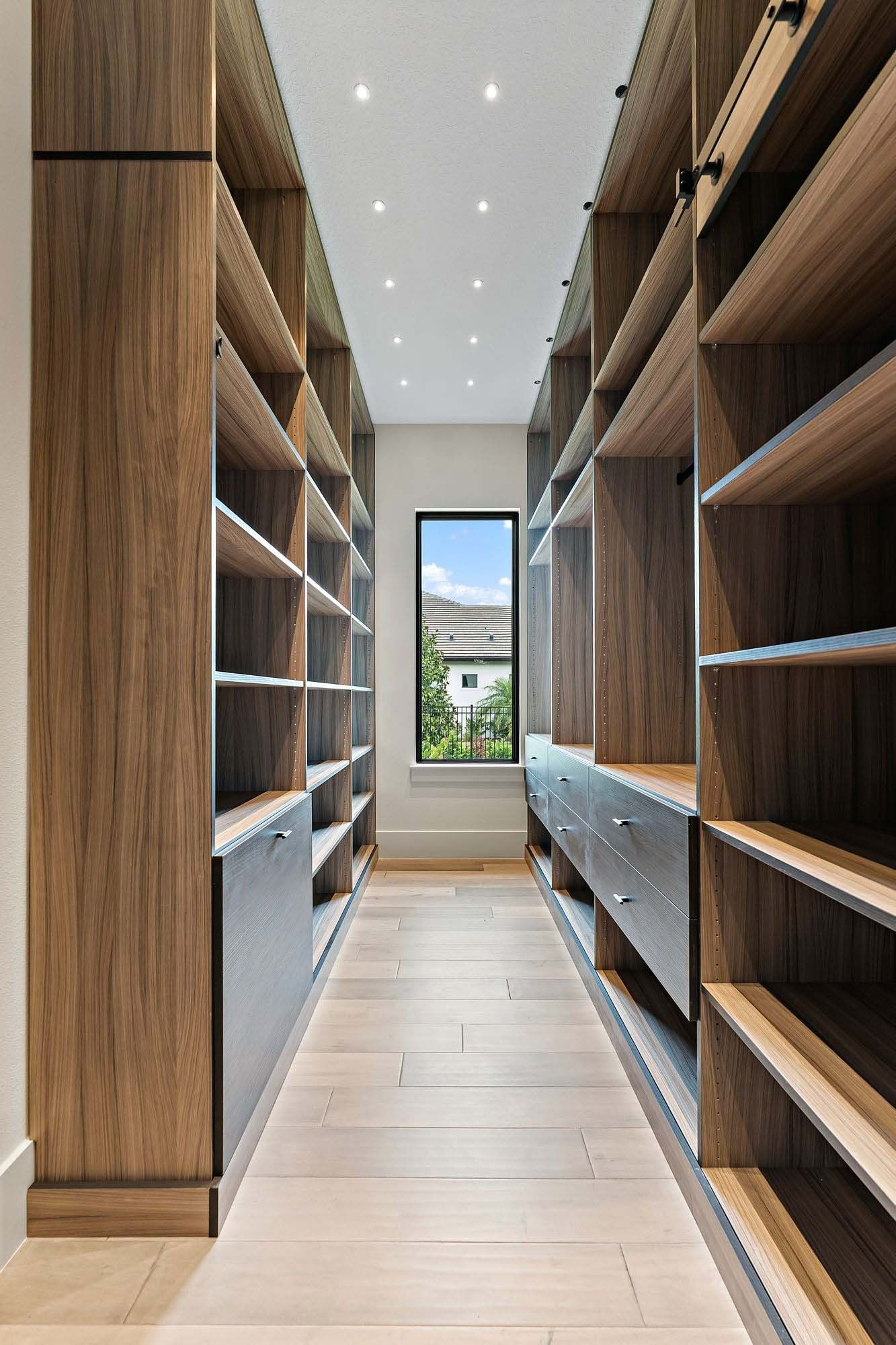 Narrow walk-in closet with wooden shelves and drawers, light floor, and a window at the end.