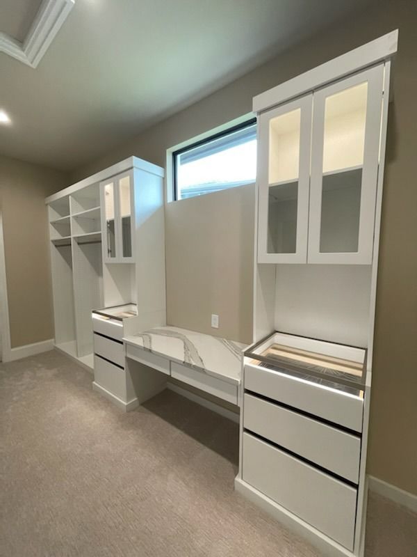 Built-in white closet shelving and drawers in a beige carpeted room with a small window.