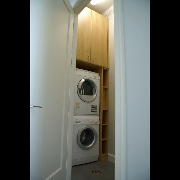 Stacked washer and dryer in a narrow laundry closet with wooden cabinets and shelving