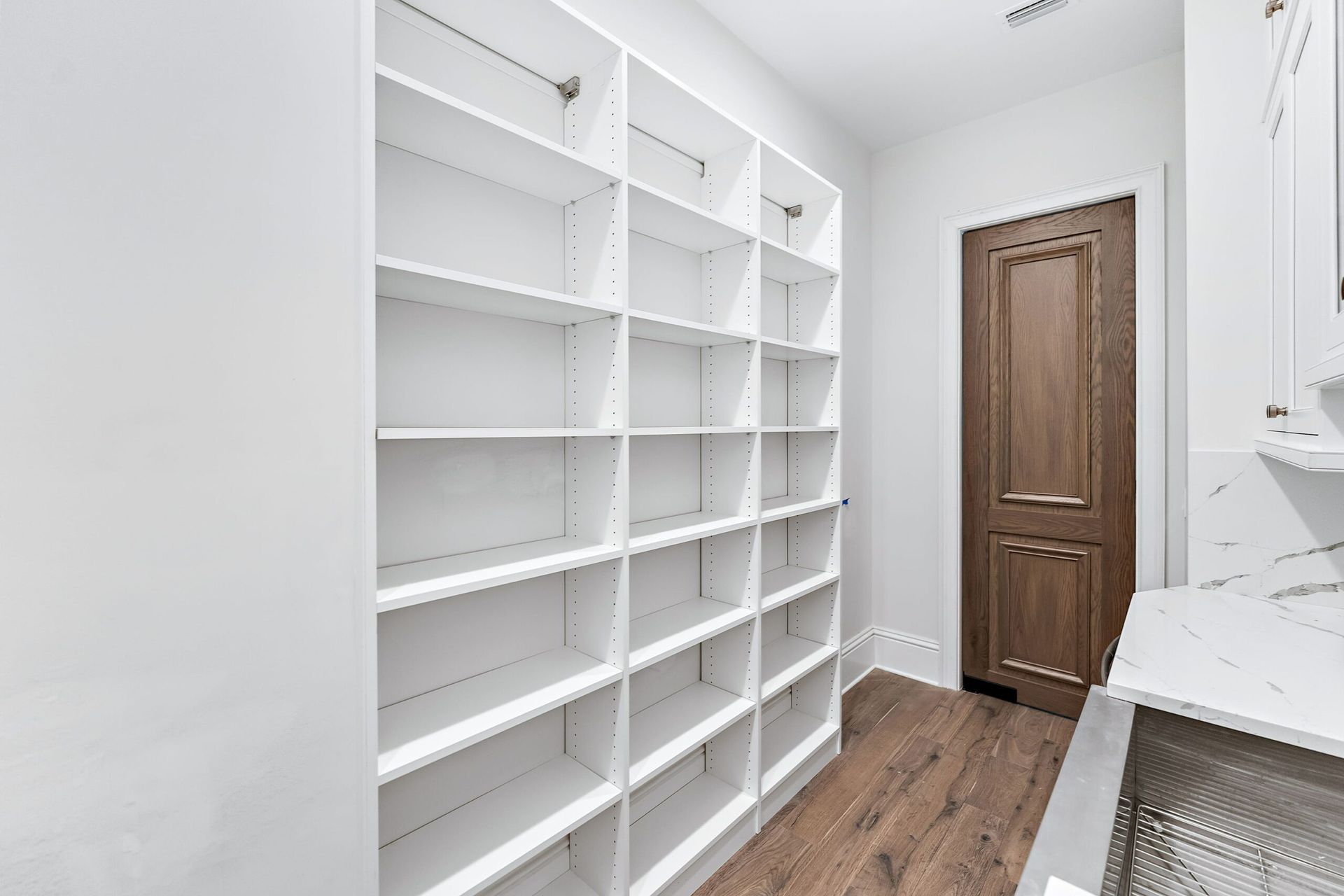 Bright white walk-in closet with empty shelves, wood floor, and a brown door at the end.