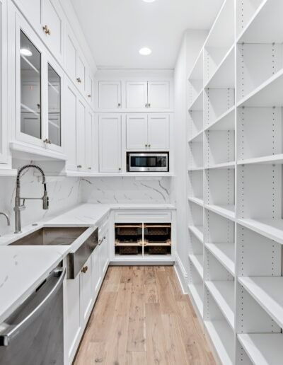 Bright white walk-in pantry with built-in shelves, counter, sink, and microwave, accented by light wood flooring.