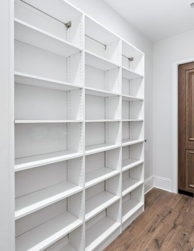 White built-in shelving in a narrow closet with wood flooring and a closed brown door.