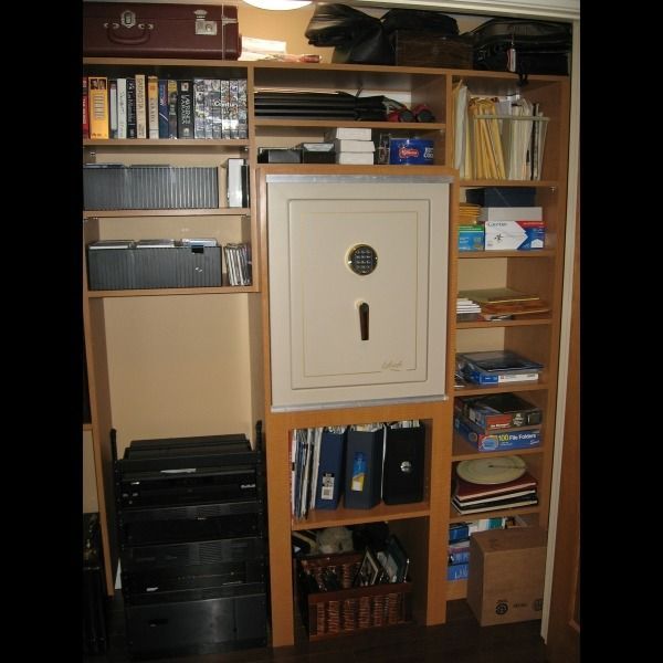 Wooden shelving unit with books, storage boxes, a wall safe, and electronics in a home office.