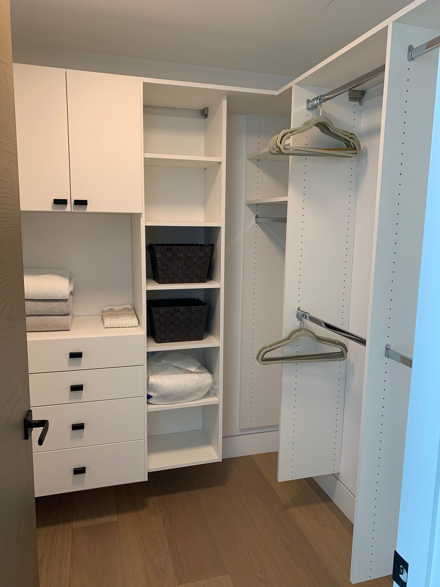 Open walk-in closet with white shelves, hanging rods, drawers, and brown wood flooring.