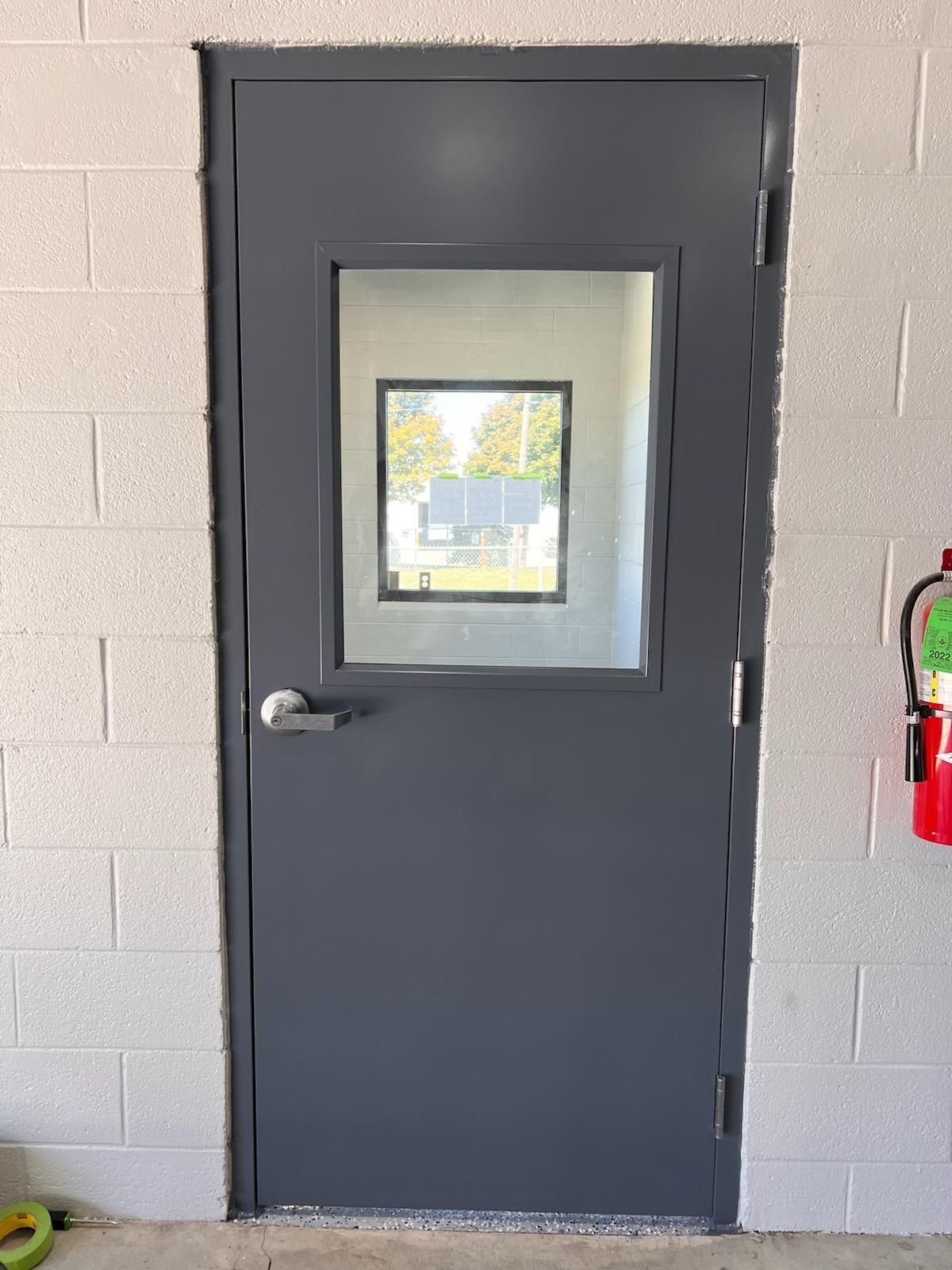A gray door with a small window in it is on a brick wall next to a fire extinguisher.