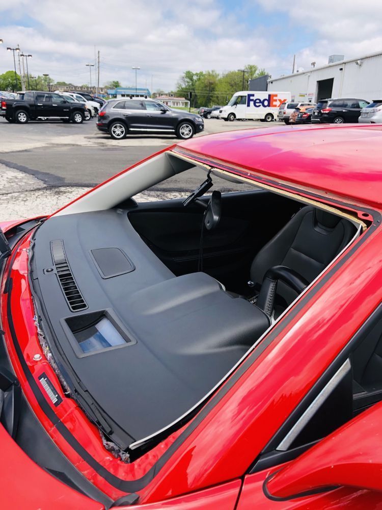 A red car with a broken windshield is parked in a parking lot with a fedex truck in the background.