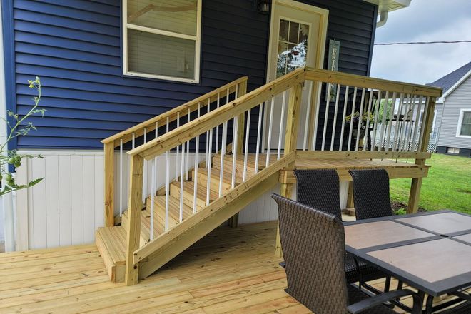 Wooden ramp with railing leading to a blue house, beside a patio table and chairs in a backyard.