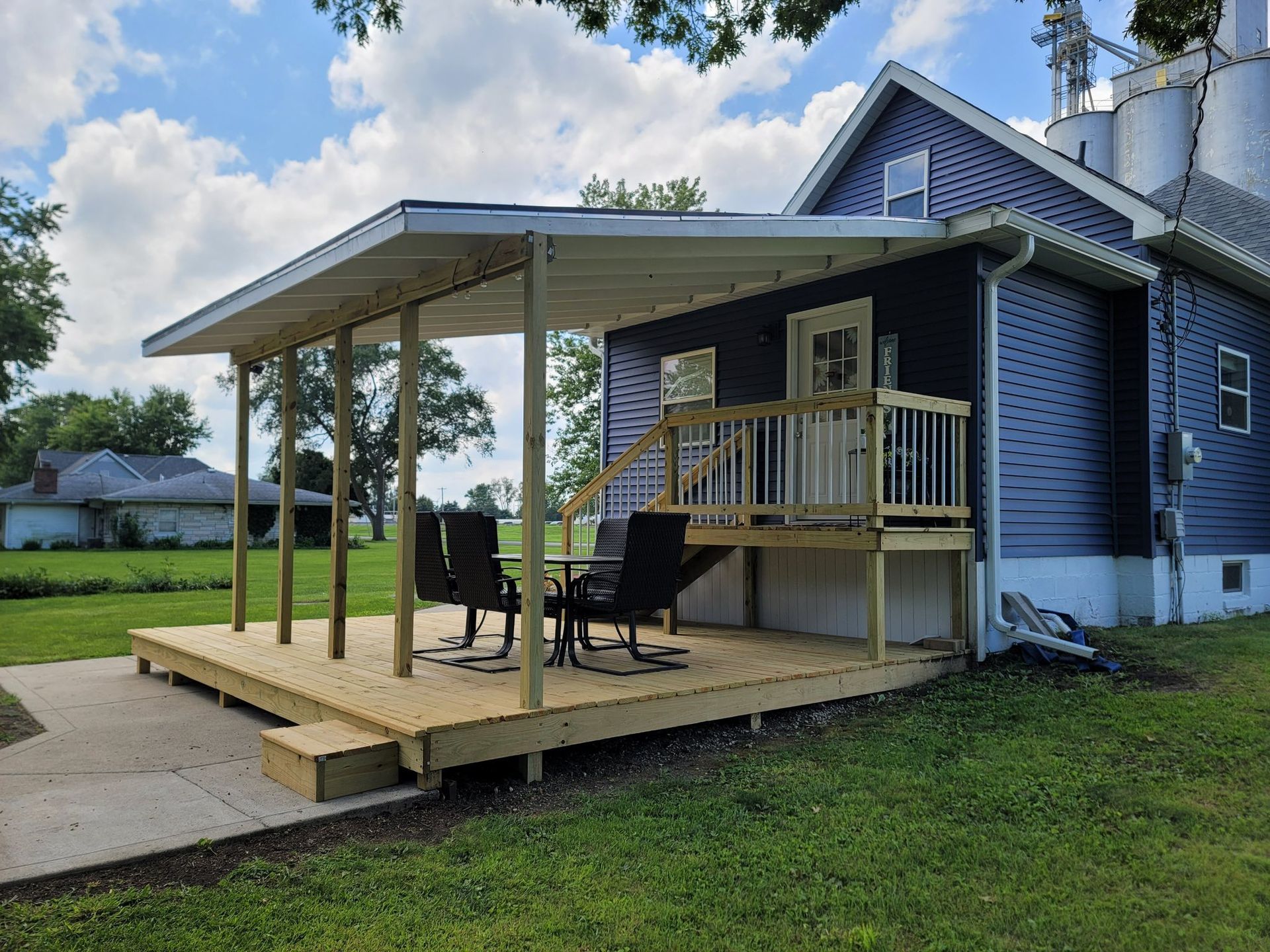 Small blue house with a covered wooden deck, chairs, and a grassy yard under a partly cloudy sky