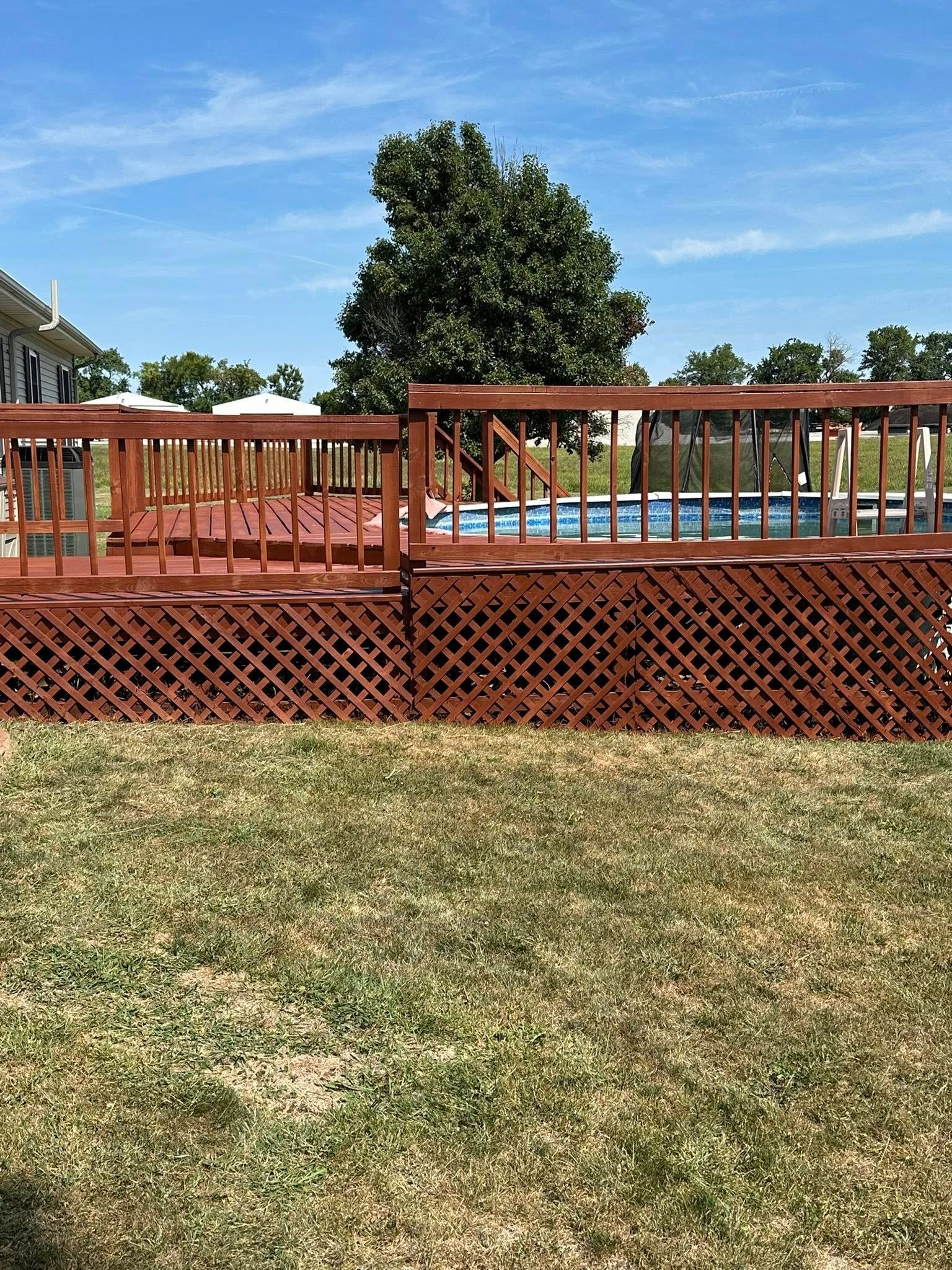 Red wooden deck with lattice, grass in foreground, and a backyard pool under a blue sky.