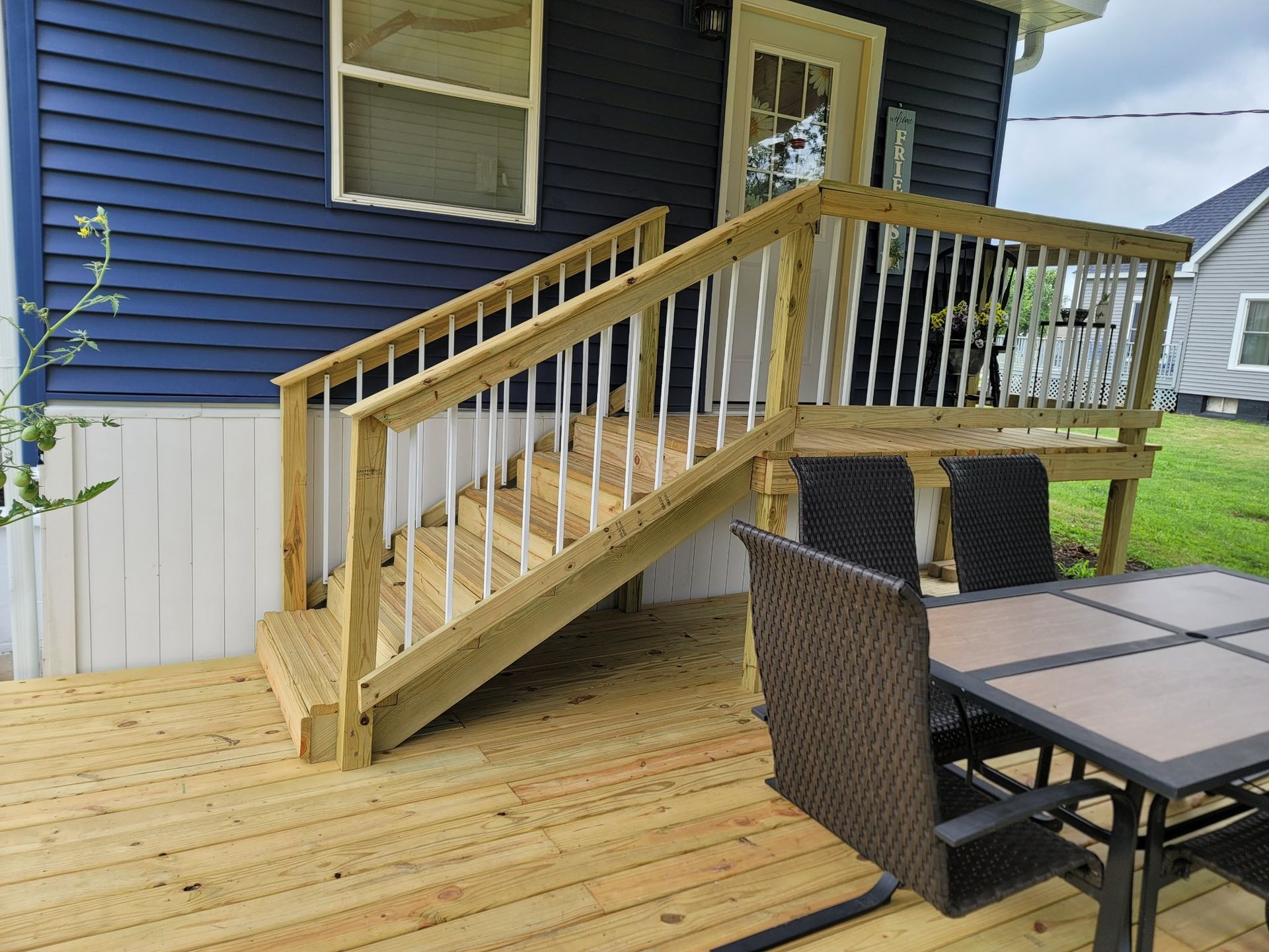 Wooden deck and ramp with railing beside a blue house, next to an outdoor table and chairs.