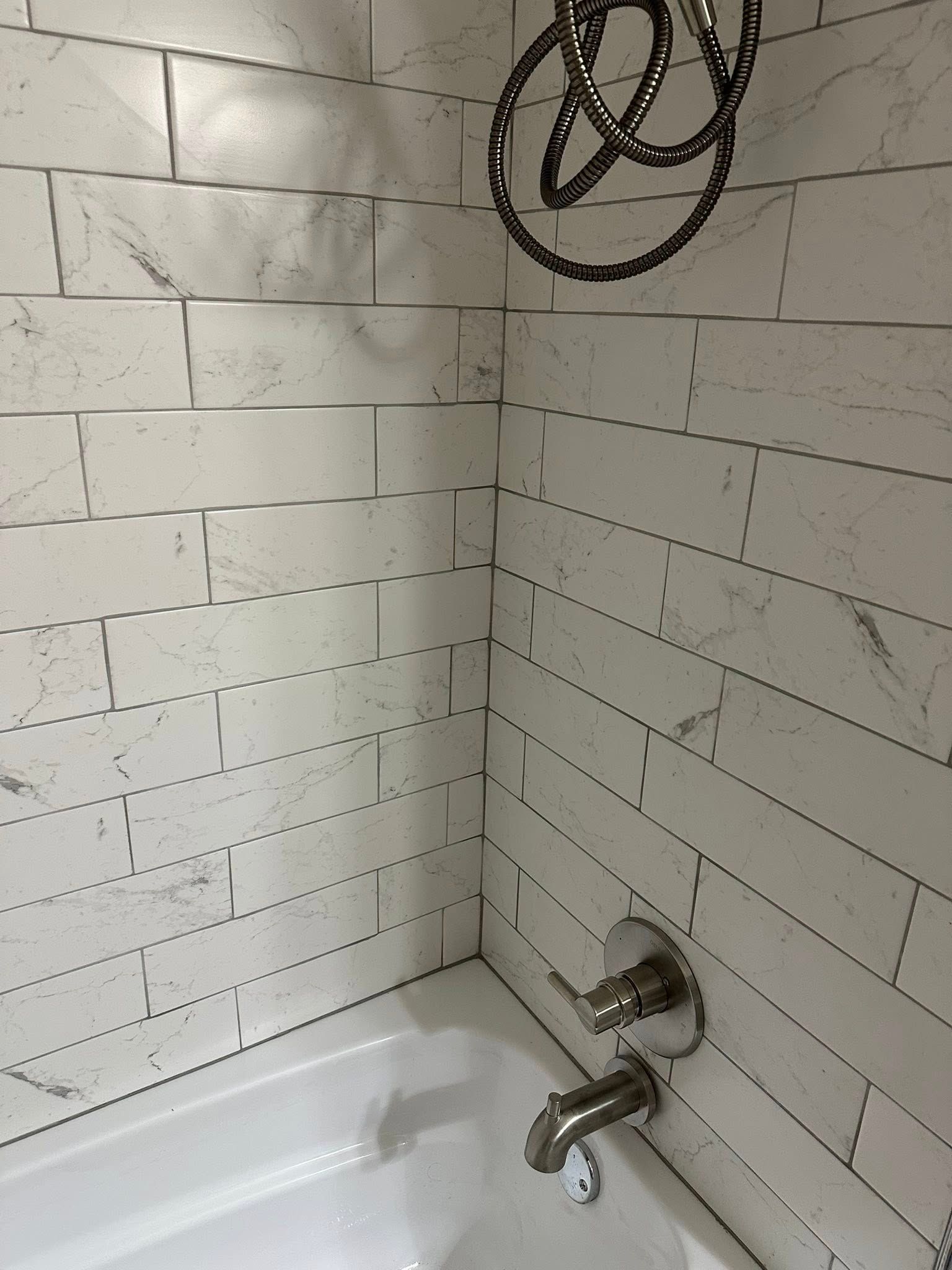 White-tiled shower corner with bathtub, chrome faucet, and shower hose hanging overhead.