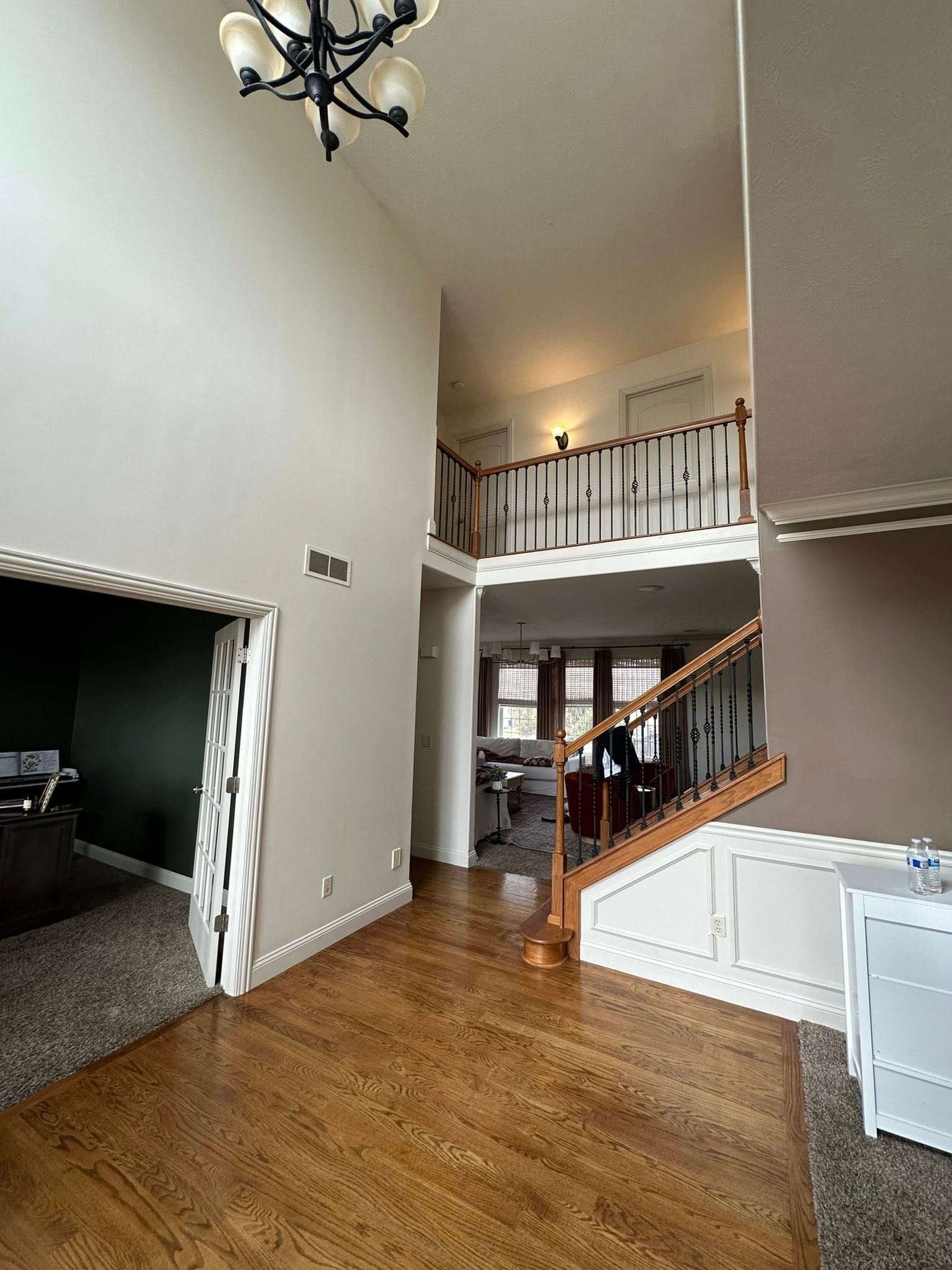 Open foyer with hardwood floors, staircase, balcony railing, and adjacent carpeted room.