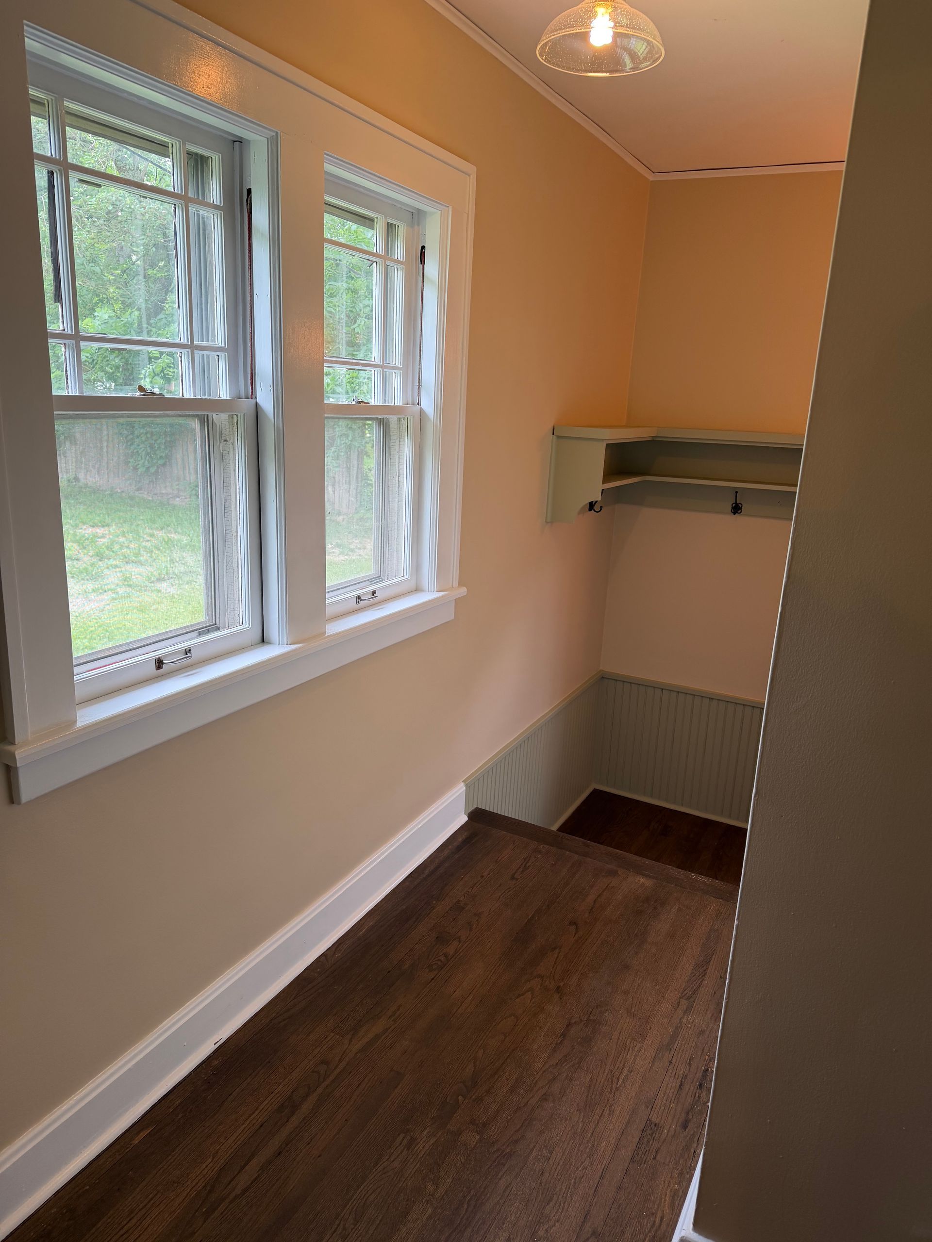 Empty small room with windows, peach walls, white trim, and a built-in shelf over dark wood floor.