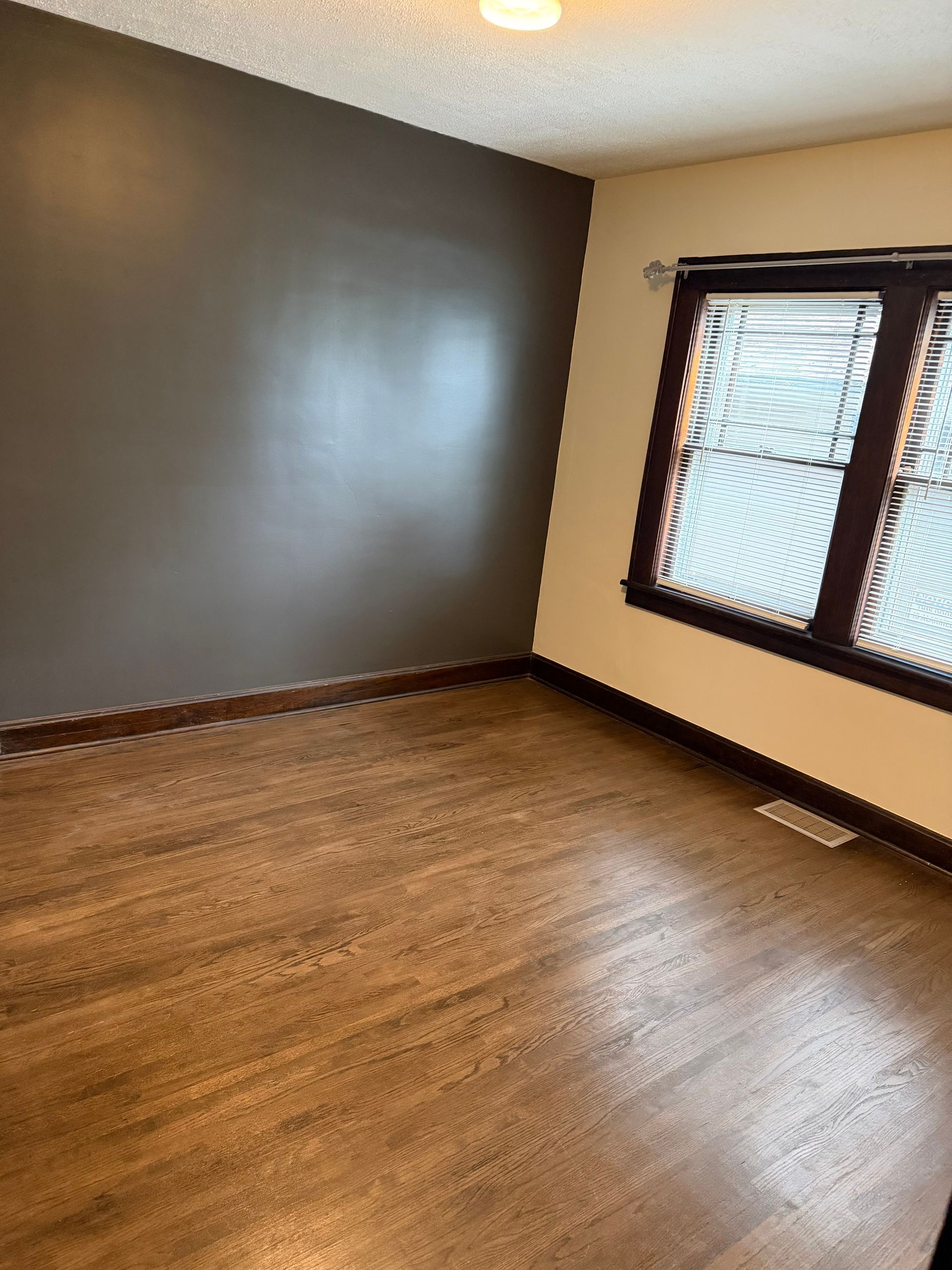 Empty room with wood floor, gray accent wall, beige walls, and a window with blinds.
