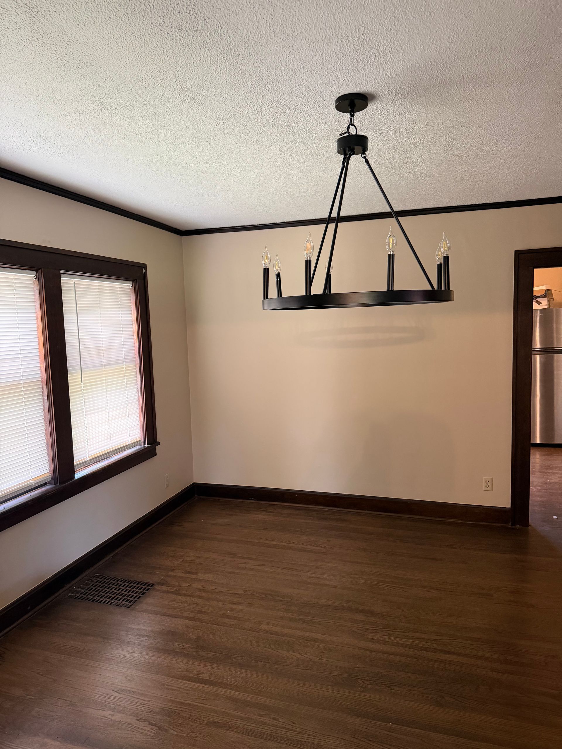 Empty room with hardwood floors, beige walls, windows with blinds, and a hanging light fixture.