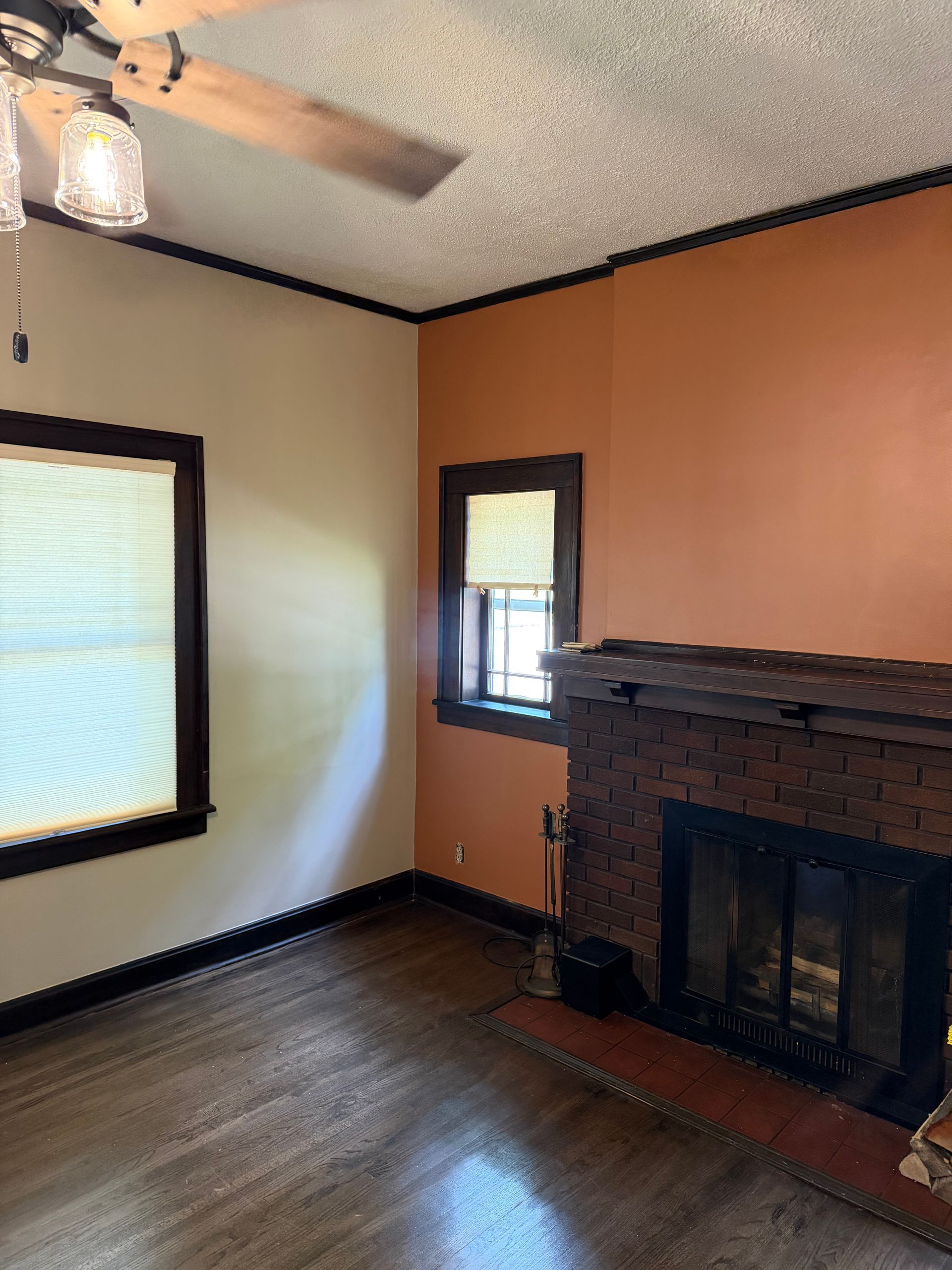 Empty living room with hardwood floors, tan walls, a brick fireplace, and two windows.