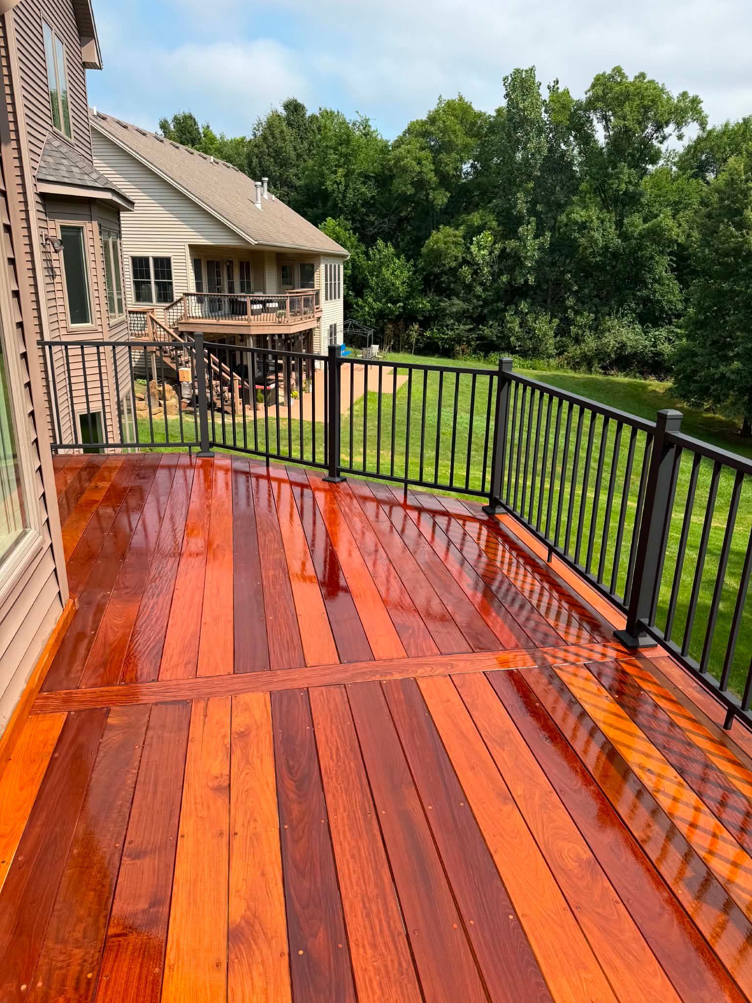 Rain-soaked red wooden deck with black railing beside a house and green trees beyond