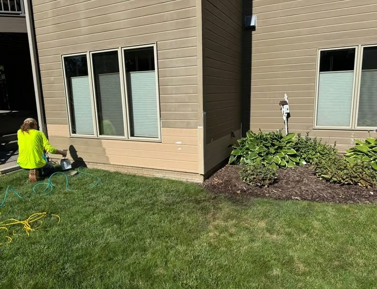Person in neon yellow shirt working near a building corner with landscaped bushes and lawn.