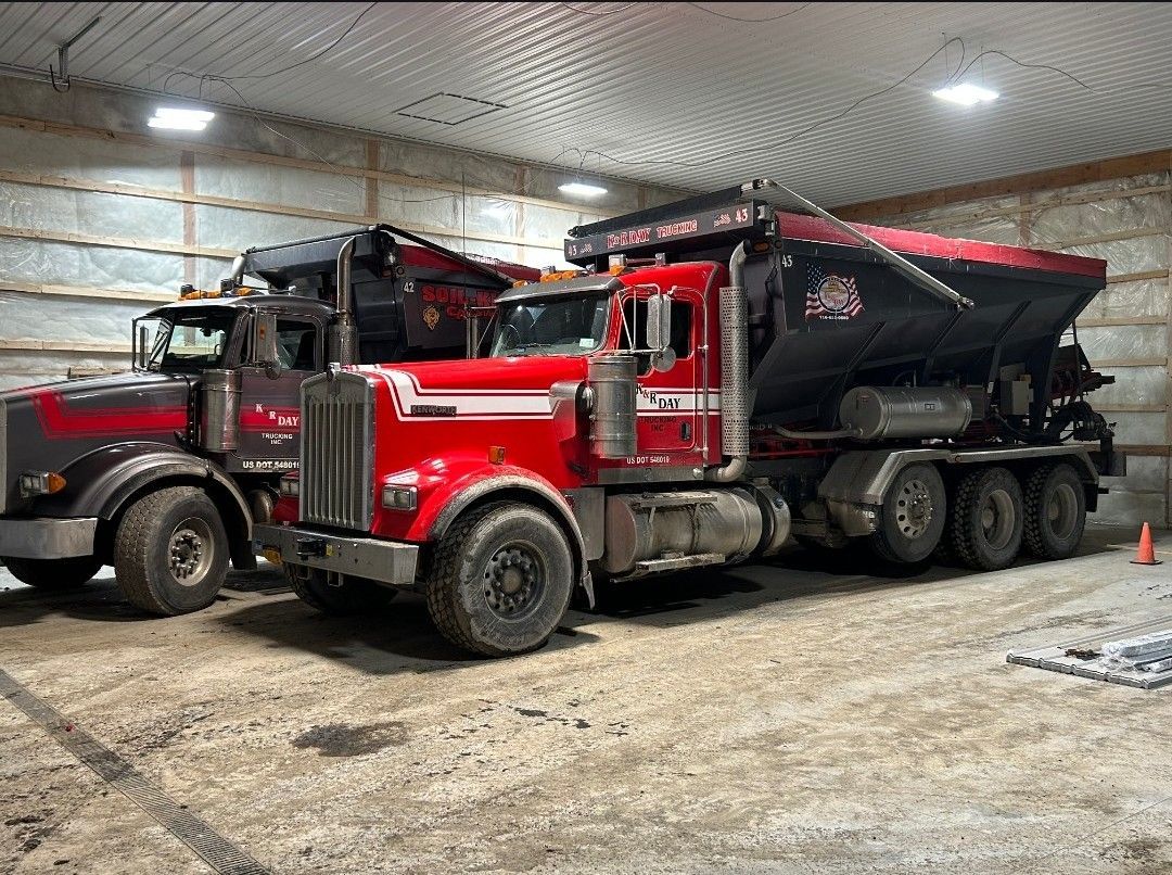 Two red and gray semi-trucks with snow plowing equipment inside a large garage.
