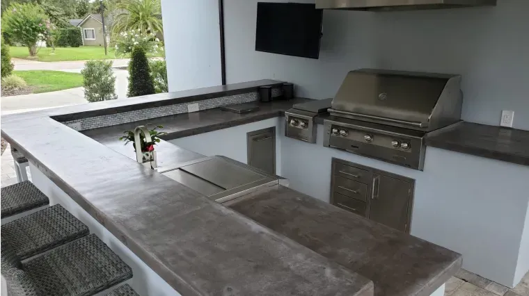 Modern outdoor kitchen with concrete bar, grill, sink, and stools under a covered patio