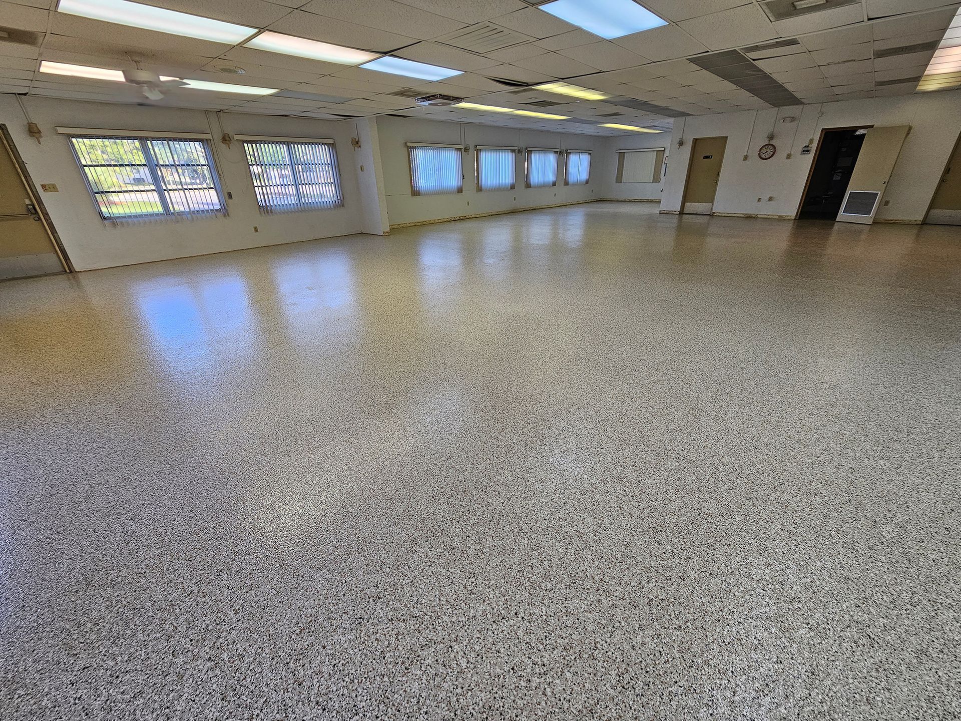 Empty fluorescent-lit room with speckled floor, windows along one wall, and a door at the back.
