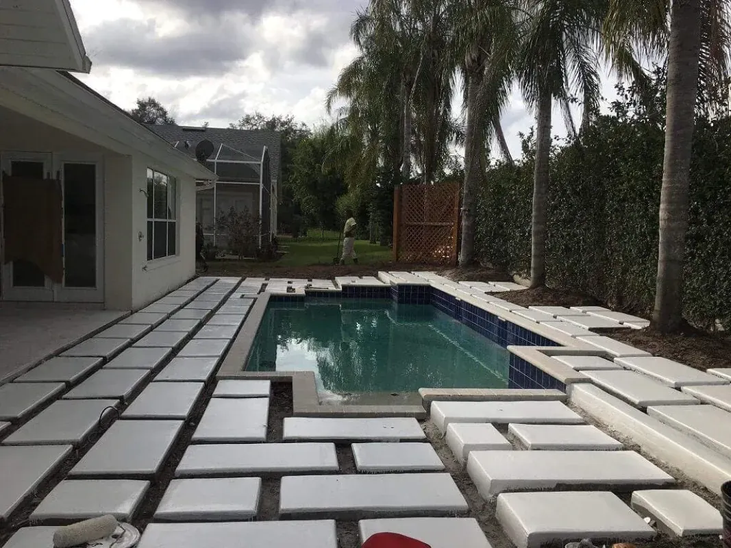 Backyard pool with white lounge cushions on a tiled patio beside a house and palm trees