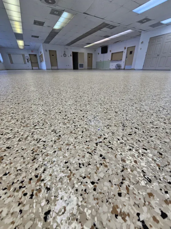 Empty speckled floor in a bright, vacant room with fluorescent ceiling lights and doors along the walls.