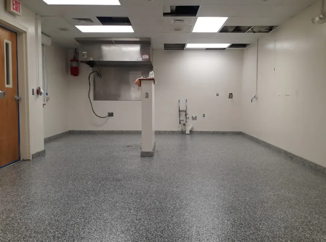 Empty white room with gray speckled floor, ceiling lights, a central support column, and a door on the left.
