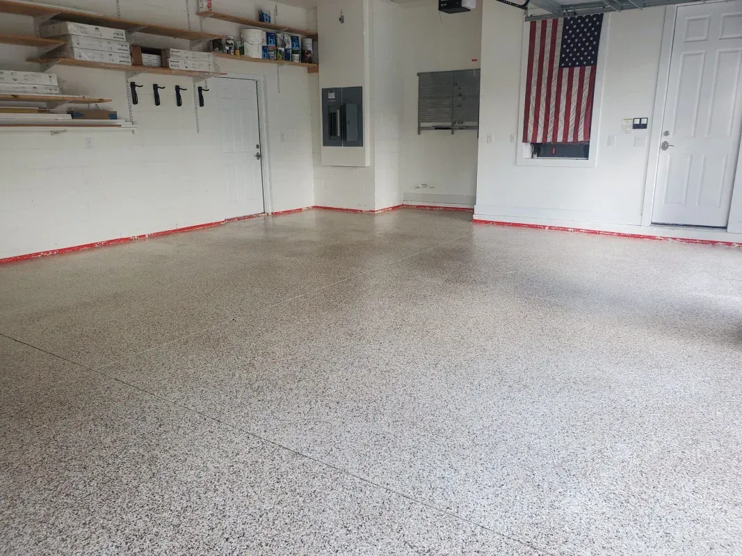 Empty garage with speckled concrete floor, white walls, shelves, and an American flag on the right.