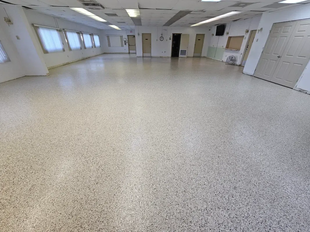 Empty bright room with white walls, windows, ceiling lights, and speckled tile floor