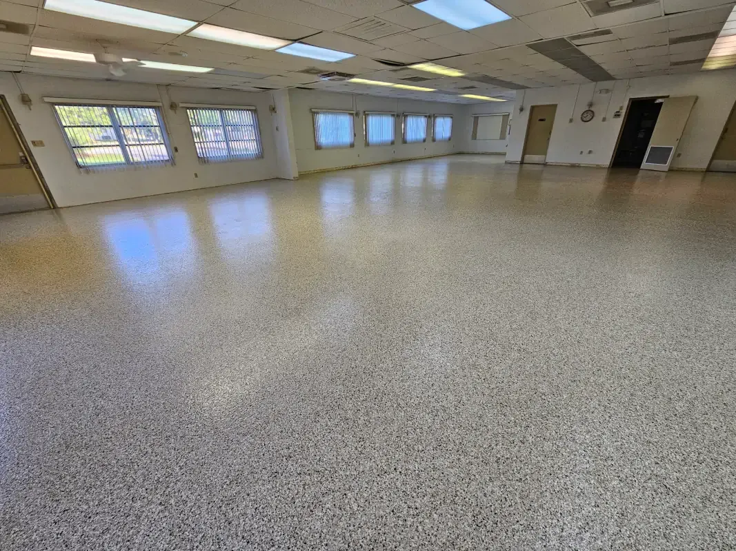 Empty bright room with speckled floor, windows, fluorescent lights, and doors along the wall
