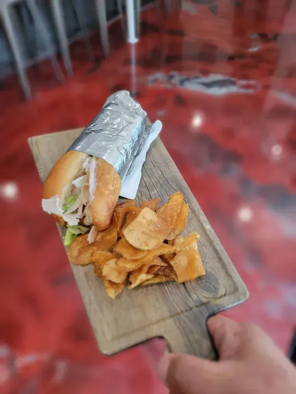 Hand holding a foil-wrapped sandwich with chips on a wooden board over a red table