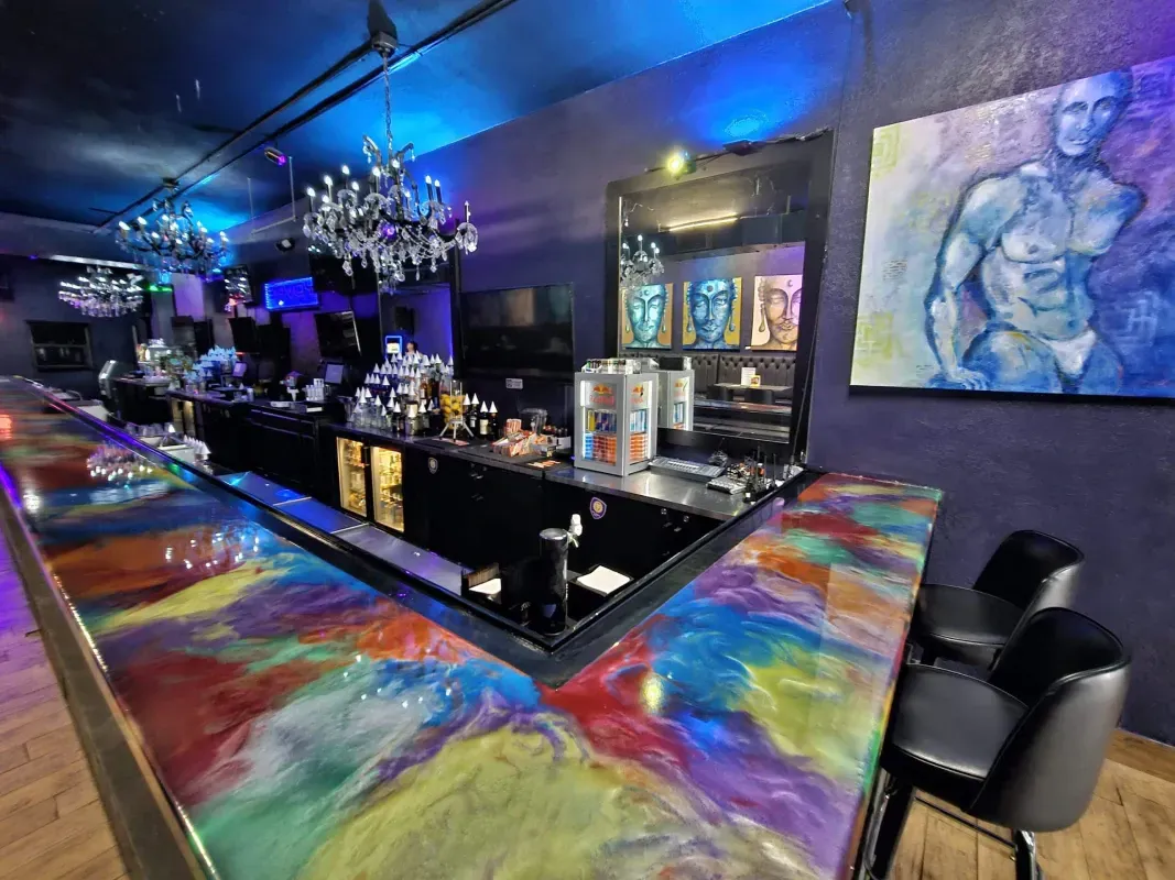 Colorful bar interior with blue lighting, mirrored counter, chandelier, and abstract wall art.