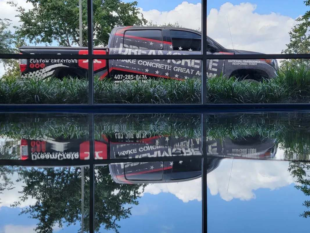 Red and black bus reflected in a glass building, with trees and blue sky in the background.