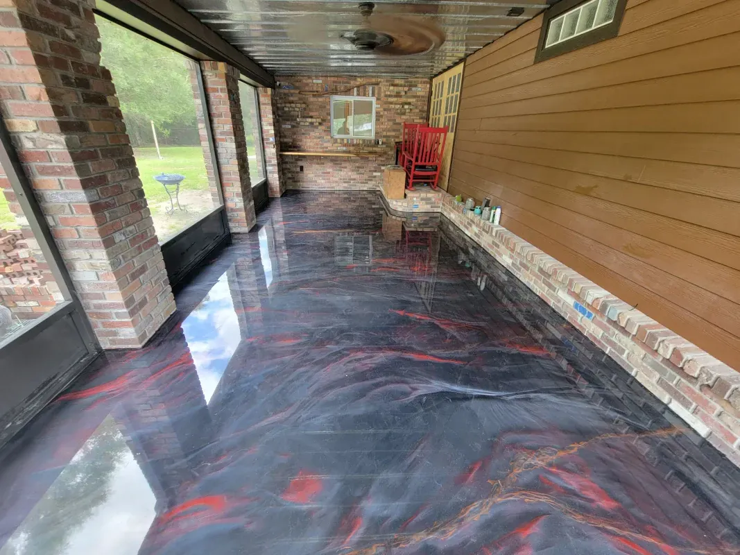 Covered porch with black plastic flooring, brick columns, tan siding, and a red chair at the far end