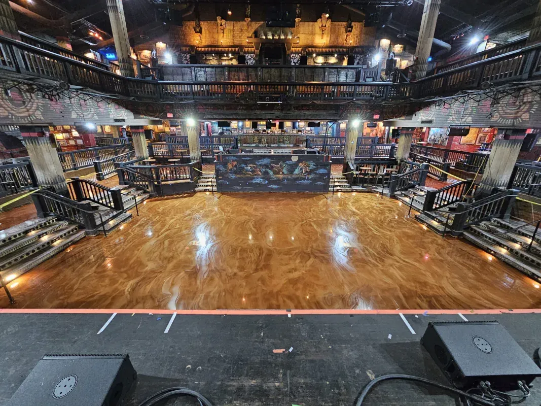 Empty concert venue with a polished wooden floor, stage lights, and seating on both sides.