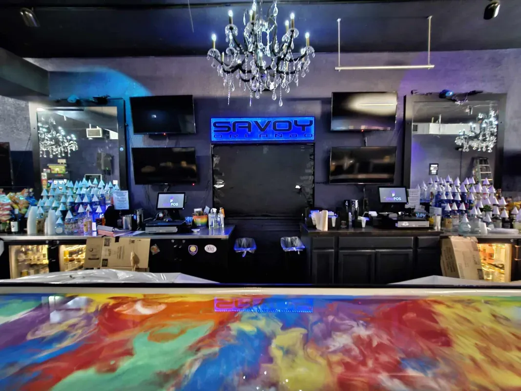 Dimly lit bar with chandeliers, multiple TVs, blue neon sign, and colorful abstract bar top.